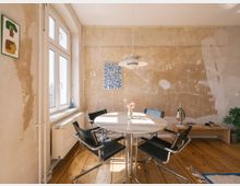 Heller Raum mit einem runden Esstisch aus Marmor, umgeben von schwarzen Stühlen mit Metallrahmen. Der Raum hat eine rohe Ästhetik mit unverputzten Wänden, Holzdielenboden, einem großen Fenster auf der linken Seite und minimaler Einrichtung wie einer Lampe, einem kleinen Regal, und Pflanzendekoration.