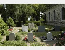 Ein gepflegter Friedhof mit mehreren Grabsteinen, die von bunten Blumen und Büschen umgeben sind. Im Hintergrund steht eine steinerne Kirche mit hohen, bogenförmigen Fenstern, eingerahmt von Bäumen und Sträuchern.