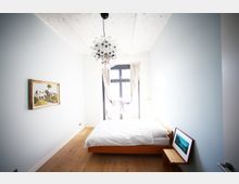 Minimalistisches Schlafzimmer mit hellem Holzboden, weiß gestrichenen Wänden und einer Decke aus weißem Backstein. Der Raum enthält ein einfaches Holzbett mit weißer Bettwäsche, ein großes Fenster mit lichtdurchlässigen Vorhängen, einen Kronleuchter aus Glas und an den Wänden ein gerahmtes Gemälde.