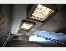 Treppenaufgang in einem U-Bahn- oder Unterführungsbereich mit Wänden aus Backstein und einer Metallverkleidung auf der rechten Seite. An der Decke befinden sich rechteckige Leuchten mit geometrischem Muster.