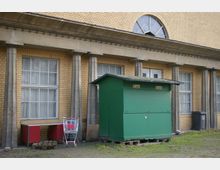 Gelbes Backsteingebäude mit großen Fenstern und angedeuteten Säulen, davor steht ein grüner Kassencontainer auf Holzpaletten und ein Einkaufswagen sowie ein rotes Möbelstück auf einem kleinen, unbefestigten Grasboden.