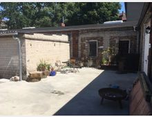 Hinterhof mit Betonboden und umgebenden gemauerten Gebäuden aus Backstein und Kalksandstein, teilweise mit vergitterten Fenstern versehen. Im Hof stehen ein Feuerkorb, mehrere Gartenstühle und Pflanzen in Töpfen, im Hintergrund Bäume und Dächer.