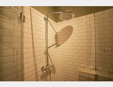 Eine Dusche mit weißen rechteckigen Wandfliesen, einem verchromten Duschkopf und einer Handbrause. Rechts ist eine Glasduschwand sowie ein weißer, wandmontierter Duschkorb zu sehen.