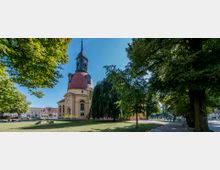 Barockkirche mit rundem Hauptgebäude und hohem Turm mit Spitzdach, umgeben von einem parkähnlichen Platz mit großen Bäumen und Rasenflächen. Im Hintergrund sind weitere Gebäude und eine Straße zu erkennen.