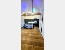 Moderne Wohnküche mit sichtbaren Holzbalken und hoher Decke, ausgestattet mit einer schwarzen Einbauküche, einer frei stehenden Kücheninsel mit Barhockern sowie einem Holzboden. Ein Oberlicht sorgt für zusätzliches Tageslicht.