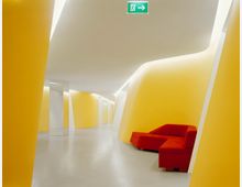 Ein moderner Innenbereich mit geschwungenen gelben Wänden, weißem Boden und minimalistischer Beleuchtung. Im Vordergrund befinden sich rote, eckige Sitzmöbel, und oben ist ein beleuchtetes Notausgangsschild sichtbar.