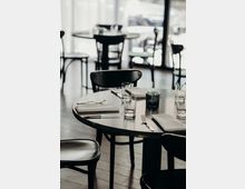 Ein modern eingerichtetes Restaurant mit runden Tischen aus dunklem Holz, die mit weißen Stoffservietten, Gläsern und Besteck gedeckt sind. Im Hintergrund sind Stühle und große Fenster zu sehen, die Tageslicht einlassen.