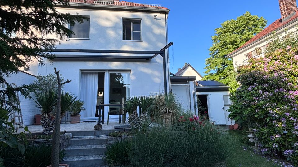Rückansicht eines zweistöckigen weißen Wohnhauses mit Flachdach, großer Schiebetür zur kleinen Terrasse und einer seitlichen Überdachung. Davor liegt ein gepflegter Garten mit Rasen, Treppenstufen, Kübelpflanzen und blühenden Sträuchern neben einem Nebengebäude.