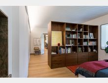 Wohnraum mit modernem Design: Ein Regal aus dunklem Holz mit offenen Fächern, gefüllt mit Büchern und Dekorationsgegenständen, steht als Raumteiler in einer hellen Wohnung mit Holzboden. Im Hintergrund ist ein weiterer Raum mit dezentem Mobiliar sichtbar.