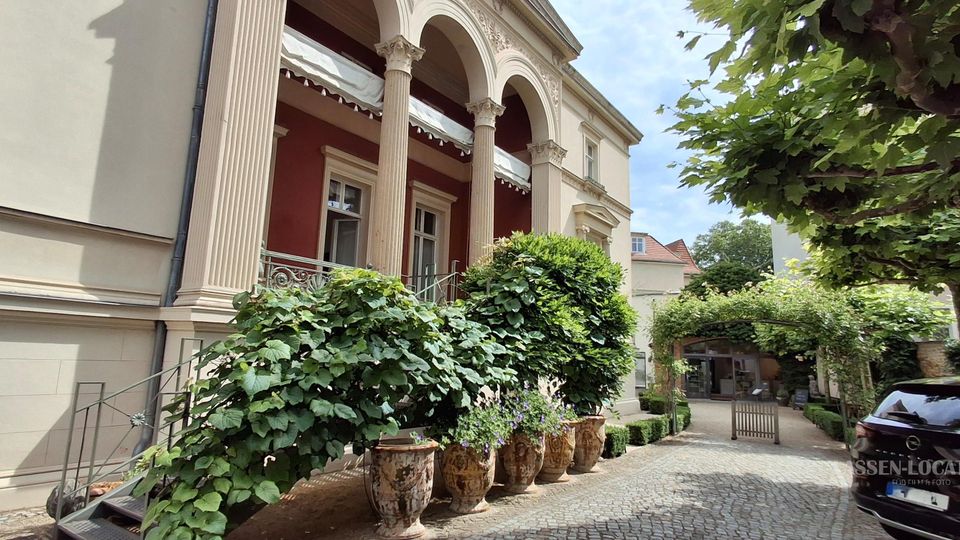 Innenhof mit Kopfsteinpflaster vor einem klassischen Gebäude mit hohen Säulen und Rundbogen-Arkaden. Große Pflanzkübel mit dichtem Grün säumen den Weg, der unter einer bewachsenen Pergola zu einem Tor und einem Nebengebäude führt.