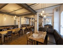 Ein gemütlich eingerichteter Speiseraum mit langen Holztischen, verschiedenen Stühlen und schwarzen Lederbänken. Die Decke ist mit Balken dekoriert, und von der Decke hängen mehrere glühende Lampen herab.