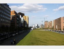 Städtischer Bereich mit modernen Gebäuden auf beiden Seiten einer breiten Grünfläche; links Gehwege und parkende Autos, rechts eine Reihe von Bäumen und Straßenlaternen, im Hintergrund Baukräne und weitere Gebäude.