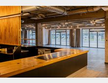 Innenraum eines modernen Cafés oder Restaurants mit einem Tresen aus Holz, sichtbaren Lüftungsrohren an der Decke und großen Glasfassaden, die den Blick nach draußen ermöglichen. Die Gestaltung kombiniert Beton- und Holzelemente und wirkt offen und industriell.