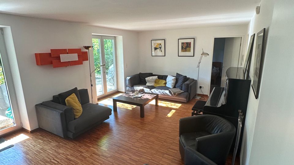 Helles Wohnzimmer mit Holzparkett, grauen Sofas und zwei schwarzen Sesseln um einen niedrigen Couchtisch. Große Fenster- und Glastüren lassen Tageslicht herein; an den Wänden hängen Bilder, daneben stehen Stehlampen und ein langes Sideboard.