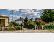 Vorgarten mit dichtem Grün und Blumen, eingefasst von einem schmiedeeisernen Tor und gemauerten Pfeilern, dahinter mehrere bunte Einfamilienhäuser und ein Strommast unter wolkigem Himmel.