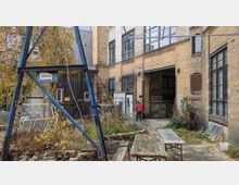 Hinterhof mit industriellem Charakter, geprägt von Backsteinwänden, großen Fenstern und einem rostigen Tor. Im Vordergrund stehen eine hölzerne Sitzbank, ein Metallgestell und wild wachsende Pflanzen, während im Hintergrund eine Person zu einem Gebäudeeingang geht.