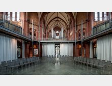 Innenraum einer historischen Kirche mit rotem Backstein und neugotischen Bögen. Der Raum ist modern umgestaltet, mit Stuhlreihen im Halbkreis, weißen Vorhängen und einer Orgel auf einer Empore im Hintergrund.