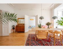 Wohnzimmer im Altbaustil mit hohen Decken, verzierten Stuckelementen und Fischgrätparkett. Ein Esstisch mit Stühlen steht auf einem roten Orientteppich, im Hintergrund ist eine Holzkommode mit vergoldetem Spiegel, Zimmerpflanzen und eine offene Doppeltür zu einem Sofa sichtbar.