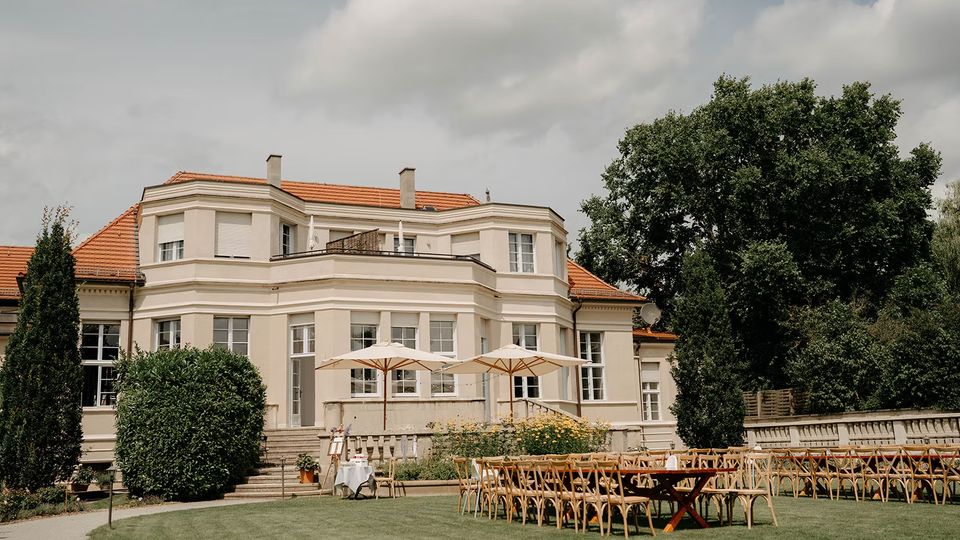 Helles, zweigeschossiges Herrenhaus mit rotem Ziegeldach, großen Sprossenfenstern und Balkonterrasse, davor eine erhöhte Steinterrasse mit Sonnenschirmen. Im Vordergrund liegt ein gepflegter Rasen mit aufgestellten Stuhlreihen und Tischen, umgeben von Büschen und großen Bäumen.