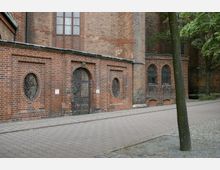 Ziegelsteinfassade eines historischen Gebäudes mit verzierten, ovalen Gittern und einem kunstvoll gestalteten Eingangstor. Rechts sind spitzbogenförmige Fenster zu sehen, und ein Baum steht im Vordergrund neben einem gepflasterten Gehweg.