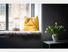 Gemütliche Fensternische in einem Innenraum mit großem Fenster, Fellauflage und gelbem Kissen; auf der Fensterbank steht ein geflochtener Korb. Daneben ein kleiner weißer Tisch mit Vase voller rosa Tulpen vor einer hellen Wand, draußen sind Backsteinfassaden zu sehen.