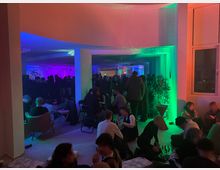 Ein großer, offener Raum mit neonfarbenen Lichtern in Blau, Grün und Pink, gefüllt mit Menschen, die in Gruppen auf Stühlen oder am Boden sitzen. Rechts ist ein Fenster und eine große Topfpflanze zu sehen, im Hintergrund eine dicht gedrängte Menge vor einer Wand mit leuchtendem Schriftzug.