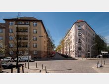 Straßenkreuzung in einem Wohngebiet mit mehrstöckigen Wohnhäusern aus hellem Putz und Backstein, Bäumen entlang der Straße und parkenden Autos. Der Straßenbelag besteht aus Kopfsteinpflaster, und Straßenschilder markieren die Kreuzung.