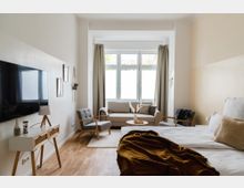Helles Zimmer mit einem großen Fenster, einem Doppelbett, einer beigen Couch, zwei grauen Sesseln und einem kleinen runden Tisch. An der linken Wand ein Flachbildfernseher über einer schmalen Kommode, Holzboden und neutrale Einrichtung mit minimalistischen Dekorationen.