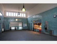 Innenansicht einer U-Bahn-Station mit türkisfarbenen Fliesenwänden, einer Uhr und Pendelleuchten an der Decke. Auf der rechten Seite befindet sich ein Bäckereistand mit der Aufschrift "Weizenfeld", daneben ein öffentlicher Münztelefonautomat. Ein Treppenabgang mit Rolltreppe ist in der Mitte sichtbar.