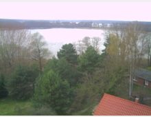 Blick auf eine baumbestandene Landschaft mit einem See im Hintergrund. Im Vordergrund ist ein Haus mit rotem Ziegeldach zu sehen.