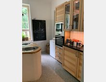 Kleine Küche mit weißen Wandfliesen, einer Arbeitsplatte aus Holz, einem eingelassenen Spülbecken und einer Einbau-Mikrowelle. Links steht ein Fenster mit Blick ins Grüne, daneben befindet sich eine schwarze Tafel mit einer Zeichnung.