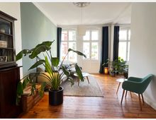 Wohnzimmer mit Holzdielenboden, großen weißen Doppelfenstern und hellen Wänden. Im Raum stehen ein grün gepolsterter Stuhl, ein Sofa, ein Teppich und mehrere Zimmerpflanzen, darunter eine große im Vordergrund. Gardinen und Möbel sorgen für eine gemütliche Atmosphäre.