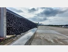 Ein Hafengelände mit einer großen Kohlehalde an der linken Seite, flankiert von einer Betonmauer. Im Hintergrund sind Kräne und ein Schiff zu sehen, während sich der Himmel wolkenbedeckt zeigt.
