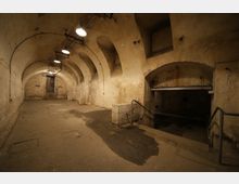 Gewölbter Kellerraum mit abgenutzten, beigefarbenen Wänden und mehreren Rundbögen, spärlich beleuchtet durch Deckenlampen. Im Vordergrund führen Treppen mit Geländern nach unten zu einem weiteren Bereich, während im Hintergrund eine Tür mit Treppen sichtbar ist.