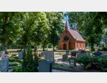 Eine kleine, rote Backsteinkapelle auf einem Friedhof, umgeben von großen Bäumen und Grabsteinen. Die Kapelle hat eine spitz zulaufende Dachform, einen Rundbogen-Eingang und kleine Bogenfenster.