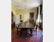 Historischer Speisesaal mit einem langen Holztisch, umgeben von geschnitzten Stühlen. Die Wände sind hellgelb gestrichen, eine Kristallleuchte hängt von der Decke, und schwere, dunkle Vorhänge rahmen die Fenster; ein bunter Orientteppich bedeckt den Holzboden.