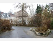 Eine kleine gepflasterte Fläche mit spärlich bewachsenen Sträuchern, einem niedrigen Steinbeet und vereinzelten Bäumen im Hintergrund. Im Umfeld sind Gebäude und eine helle Mauer sichtbar.