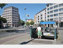 Straßenszene mit Eingang zur U-Bahn-Station "U-Bhf Blissestraße" in Berlin. Im Hintergrund sind mehrstöckige Wohn- und Geschäftsgebäude sowie eine Kreuzung mit Verkehrsampeln zu sehen.