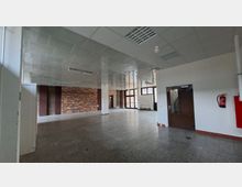 Großer, leerer Innenraum mit gefliestem Boden, abgehängter Decke mit Rasterleuchten und weißen Stützsäulen. An einer Seite befinden sich eine Ziegelwand, mehrere Türen und eine Fensterfront mit Tageslicht; rechts ist ein Feuerlöscher an der Wand montiert.
