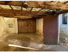 Innenansicht einer alten Garage mit Holzbalkendecke und teilweise durchsichtigem Wellplasten-Dach. Eine hölzerne Doppeltür bildet den hinteren Abschluss, während die Wände aus grobem Putz und Backstein bestehen; der Boden ist aus verdichtetem Sand oder Erde.