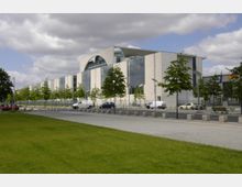 Modernes Regierungsgebäude mit heller Fassade, großen Glasfenstern und einem markanten Bogendetail im oberen Bereich, umgeben von jungen Bäumen, einer Straße mit geparkten Autos und einem Grünstreifen im Vordergrund. Der Himmel ist teils bewölkt.