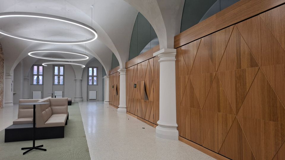 Moderner Innenraum einer Lobby oder Wartehalle mit hohen Gewölbedecken, weißen Säulen und großen ringförmigen Deckenleuchten. Rechts verläuft eine lange Holzvertäfelung mit geometrischem Muster, links stehen Sitzbänke und kleine Tische vor hohen Fenstern.