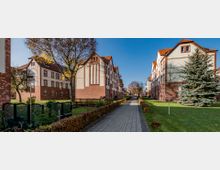 Gepflegte Wohnanlage mit mehreren Reihenhäusern aus rotem Backstein und hellem Putz, flankiert von einer gepflasterten Gehwegallee. Auf beiden Seiten befinden sich Rasenflächen, Bäume und vereinzelte Sträucher, die bei klarem, sonnigem Wetter zu sehen sind.