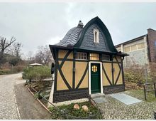 Kleines Fachwerkgebäude mit gelb verputzten Wänden, dunkelgrünen Holzbalken und geschwungenem, dunklem Schieferdach. Das Gebäude steht in einer parkähnlichen Umgebung mit bepflanzten Beeten, gepflasterten Wegen und umliegenden Bäumen.