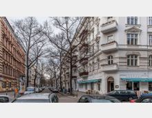 Straßenszene mit mehrstöckigen Wohnhäusern im Altbaustil, links mit roter Fassade und rechts mit weißer Putzfassade und verzierten Balkonen. Kahle Bäume säumen die Straße, auf der parkende Autos und wenige Passanten zu sehen sind.