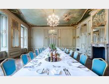 Historischer Speisesaal mit reich verzierten Wänden und Decken im Rokokostil, vergoldeten Ornamenten, einem Kronleuchter und einem Deckengemälde. Ein langer, festlich gedeckter Tisch mit blauen Stühlen steht in der Mitte des Raumes.