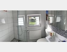 Helles, gefliestes Badezimmer mit begehbarer Glasdusche, kleinem Fenster über einem Heizkörper und einem Waschtisch mit großem Spiegel und Mosaikfliesenbordüre. Schwarz-weiß gemusterte Bodenfliesen ergänzen die weißen Wandfliesen.