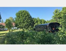 Ein ländlicher Garten mit üppigem Grün und einem großen schwarzen Bus, der teils von Vegetation umgeben ist. Im Hintergrund steht ein weißes Gebäude mit rotem Ziegeldach, flankiert von mehreren Bäumen.