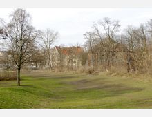 Weitläufige Grünfläche mit vereinzelten Bäumen und kahlen Sträuchern im Vordergrund, im Hintergrund mehrstöckige Gebäude mit roten Dächern und historischen Fassaden, teils von Bäumen verdeckt.