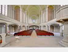 Innenraum einer historischen Kirche mit hohen, gelblich-grün gewölbten Decken, langen Holzbänken und einer zweistöckigen Empore. Große Buntglasfenster dominieren die Rückwand, ergänzt durch Säulen und verzierte architektonische Details.